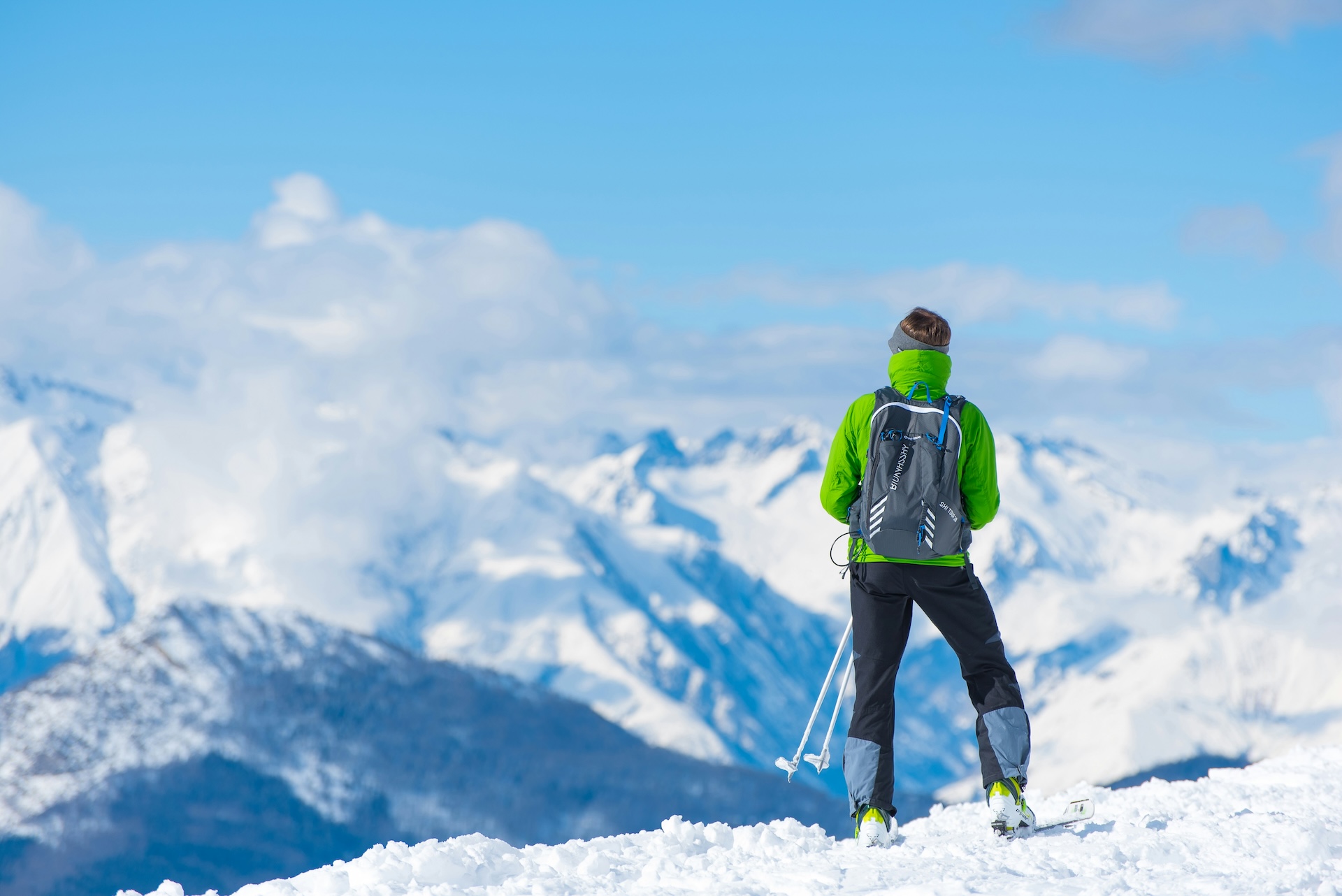 A man wearing a lime green jacket and a backpack holds hiking poles on top of a snowy mountain overlooking other peaks.