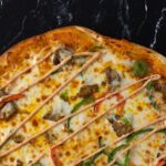 The top half of a pizza is visible in front of a black stone countertop, with cheese, veggies and drizzled orange colored sauce.