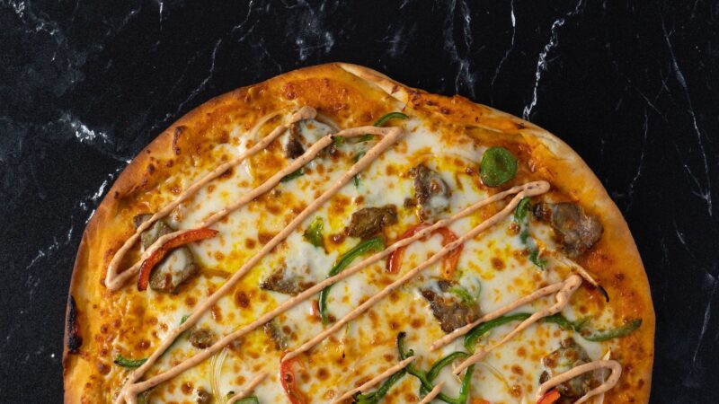 The top half of a pizza is visible in front of a black stone countertop, with cheese, veggies and drizzled orange colored sauce.