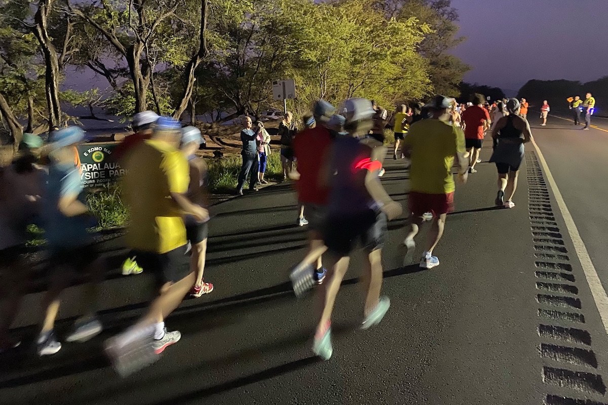 People run in a marathon on a road lined with trees and the sky is a dark hue of purple.