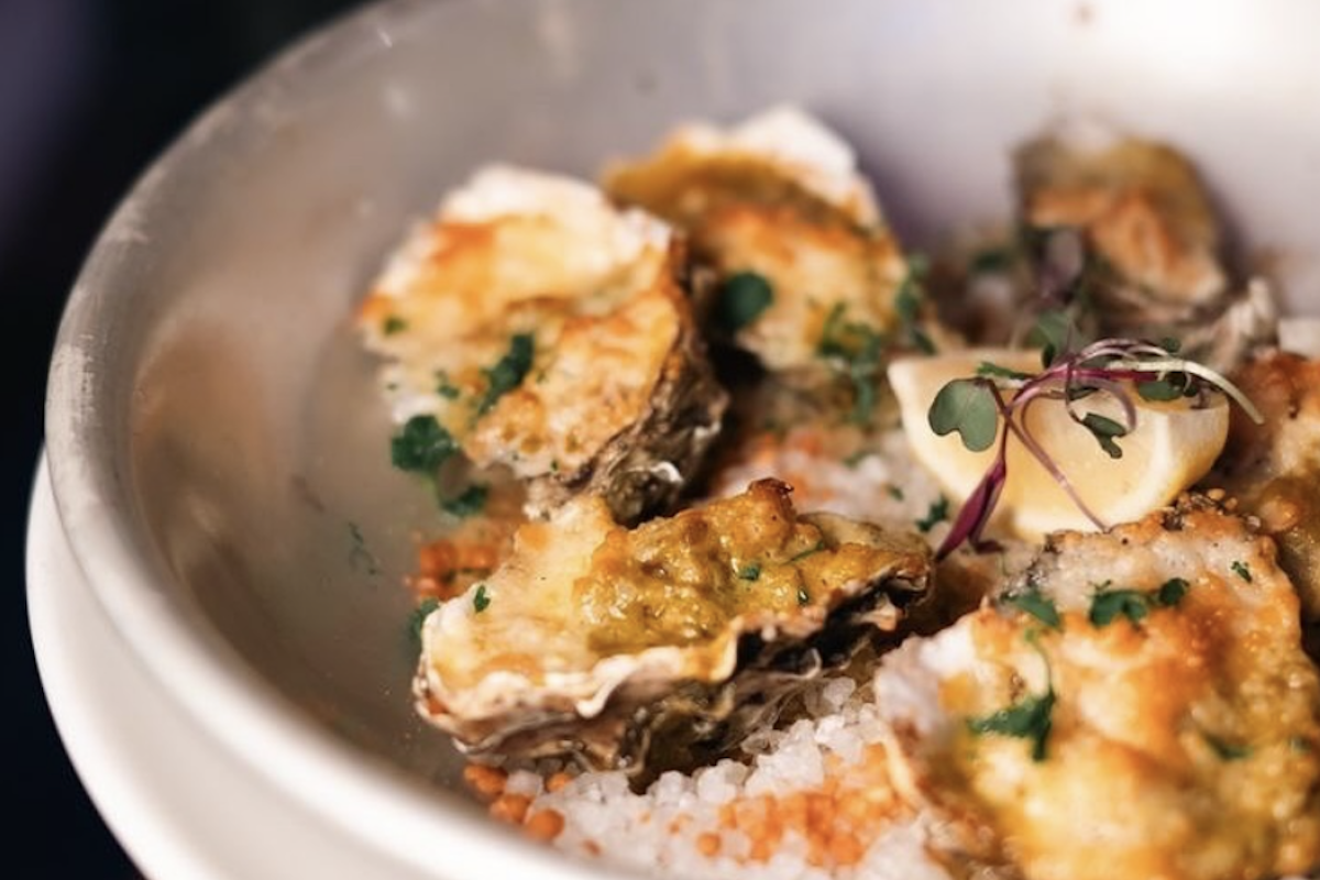 Shellfish of some kind sits atop a bed of rice, with lots of seasoning, sauce, microgreens and lemon wedges on top.