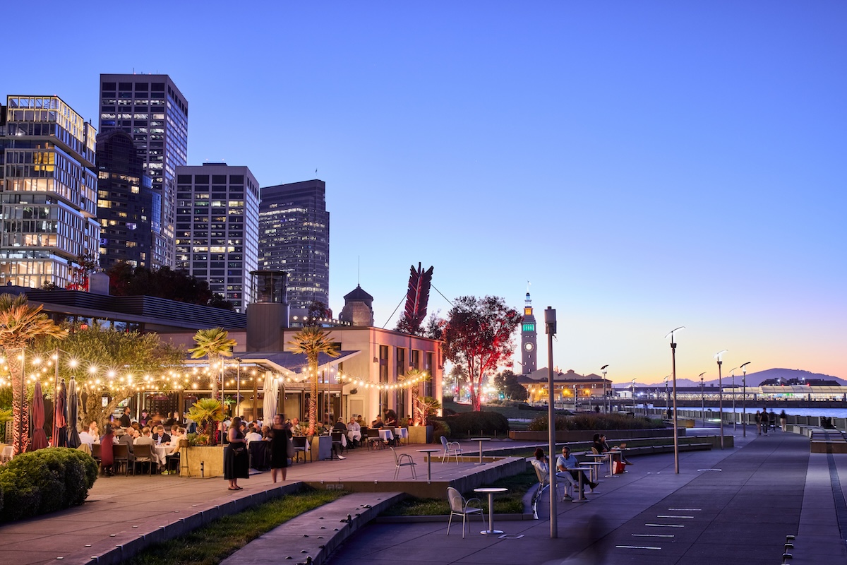 Patio seating of Epic Steak lit up at night by string lights and surrounding skyscrapers, on the Embarcadero with SF's Ferry Building in view