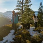 Three people snowshoe as a dog follows them, on top of a snowy mountain with trees and other mountains around them.