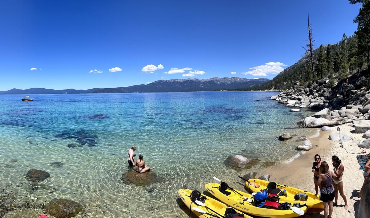 A view of Lake Tahoe from the shore, which people in swimwear stand on and yellow kayaks sit.