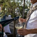 Outside, a man pours wine from a dark bottle into a wine glass held by a man wearing a white T-shirt and gray sweater tied around his neck.