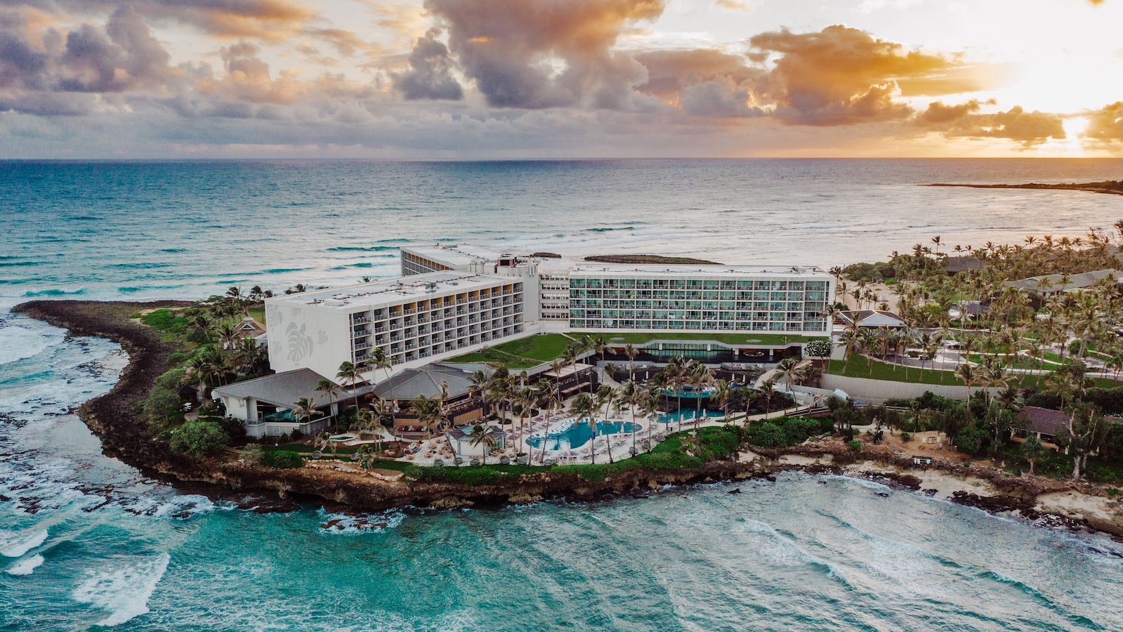 A white hotel on green land is surrounded by aqua colored water and a sunset.