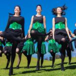 A group of women wearing black clothes, green corsets and some wearing green and blacked checkered skirts perform an Irish dance on grass in front of a bright blue sky.