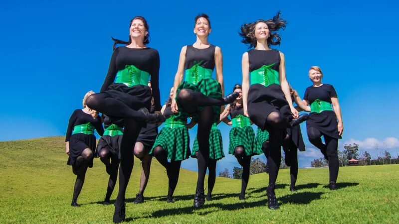 A group of women wearing black clothes, green corsets and some wearing green and blacked checkered skirts perform an Irish dance on grass in front of a bright blue sky.