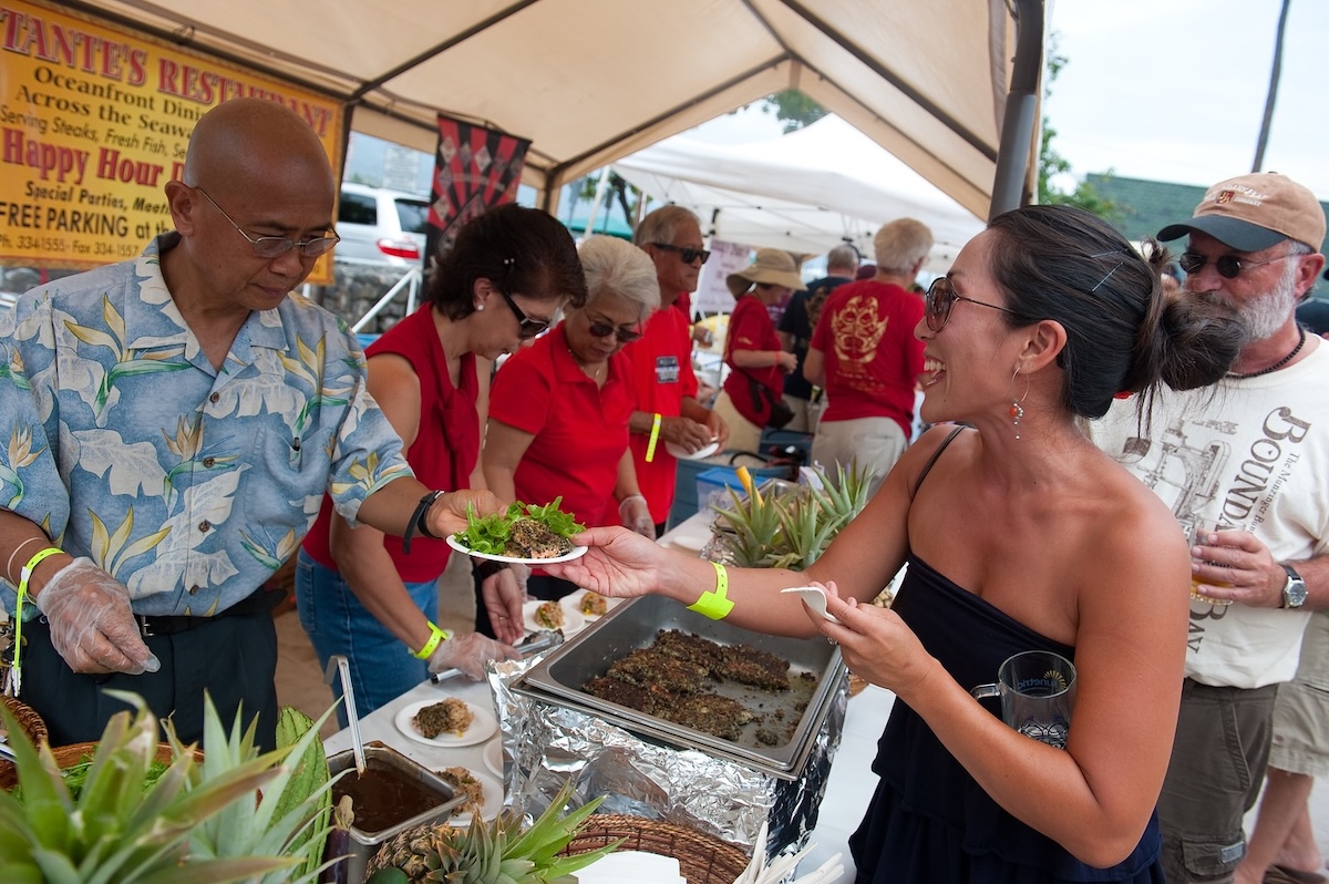 A woman receives a plate of food from a vendor in a tent with trays of food in front of him.