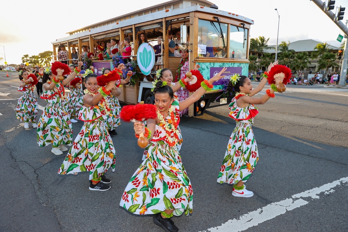 A group of women wearing patterned dresses, leis and hairpieces, holding something featured as they perform in the street. An open air bus with people is behind them.