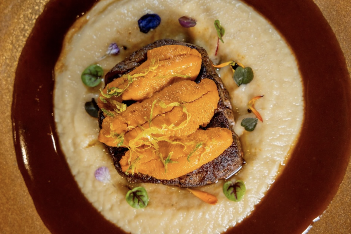 A gold plate with a bed of dark red and light tan sauces, a slab of steak, seafood and microgreens for decor.