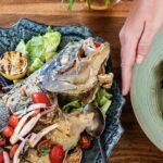 Someone holds the edge of a pale green plate with prawns and veggies, with another darker plate sits next to it with a baked fish surrounded by veggies and other toppings.