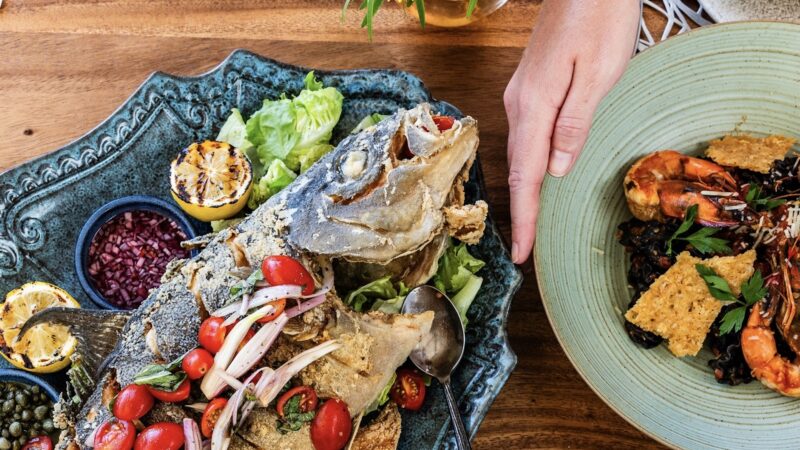 Someone holds the edge of a pale green plate with prawns and veggies, with another darker plate sits next to it with a baked fish surrounded by veggies and other toppings.