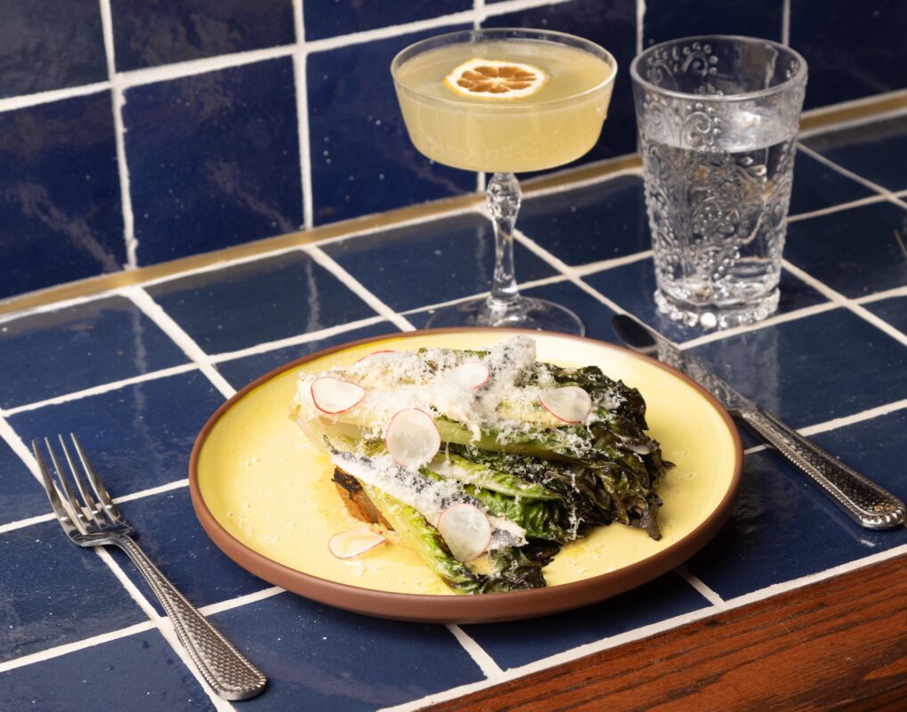 On a dark blue tiled countertop sits a butter yellow plate with cooked lettuce, sliced radish and parmesan. A fork, knife, water glass and yellow cocktail are next to the plate.