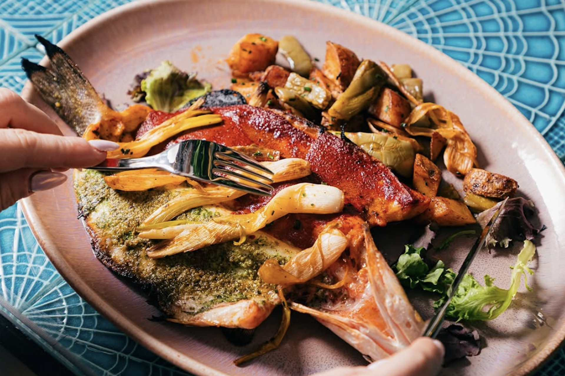 A plate of crispy fish with roasted veggies on the side, all on a pink plate atop a blue patterned table as someone cuts in.