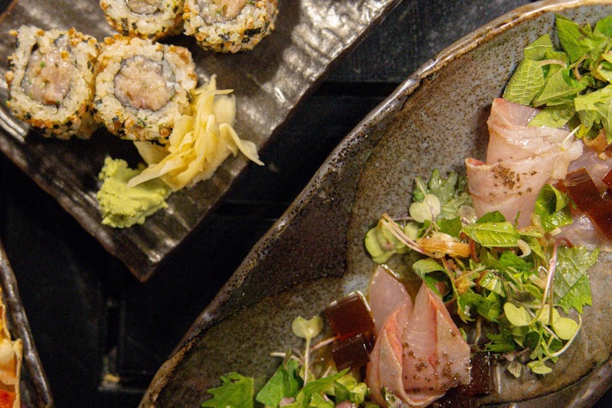 A plate with microgreens and thinly sliced sushi sits next to another plate with sushi rolls.