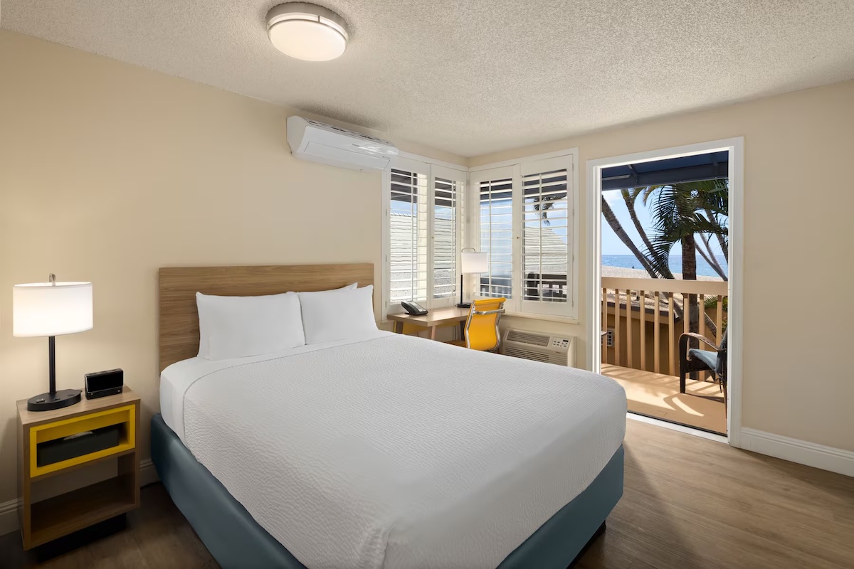 A bed with a white bedspread in a hotel room with a balcony and a view of the beach.