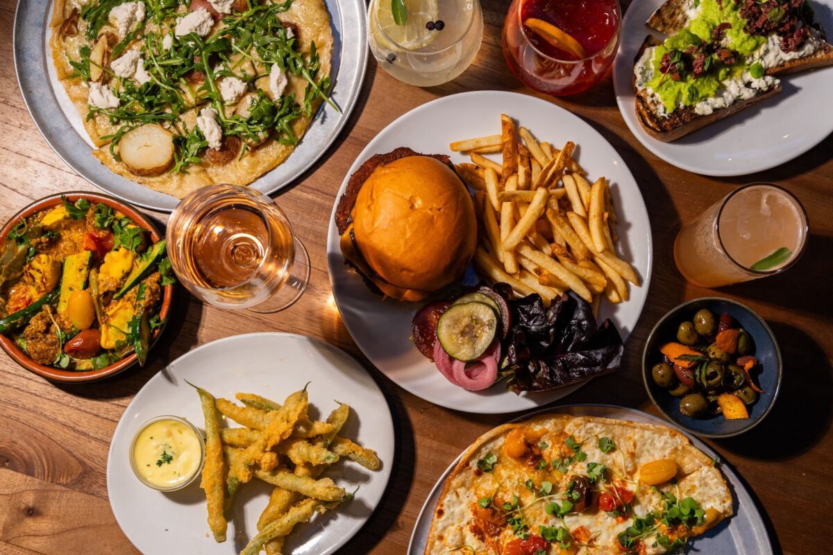 An above shot of a table with plates of food including a burger and fried, fried green beans, pizzas and more.