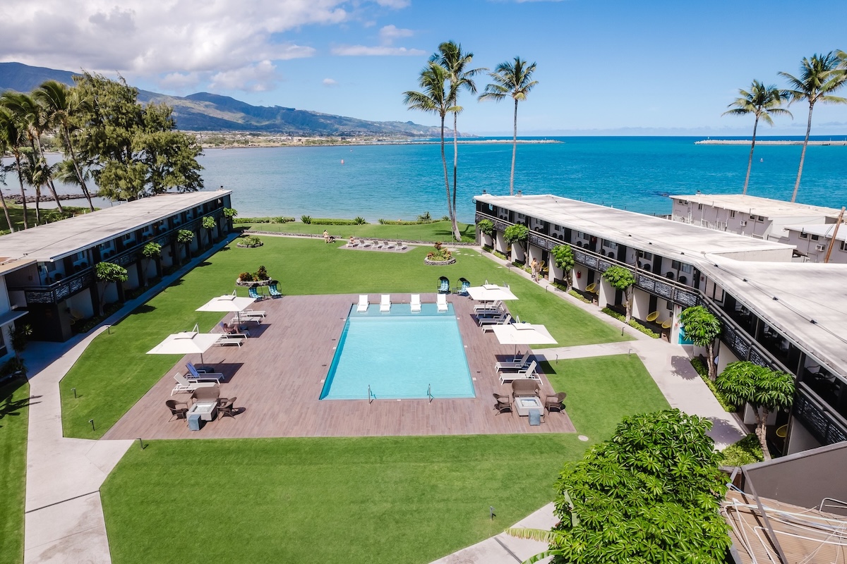 A shot overlooking a grassy area with a pool central, hotel rooms along the sides and a view of the ocean in the distance.