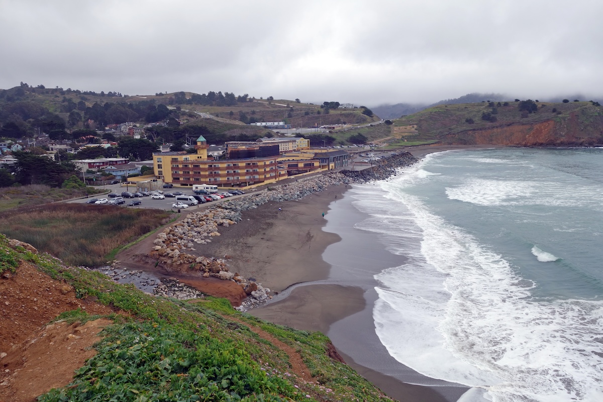 A wide shot of a beach with dark sand, rocky bluffs and buildings along the edge.