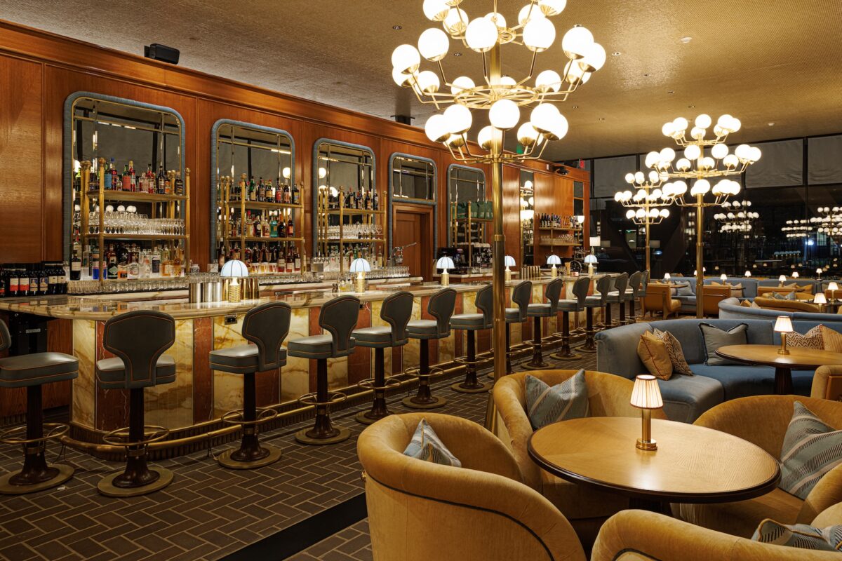 The view of a bar with gray bar stools, wood paneled walls with shelves of alcohol, large chandeliers and small tables with arm chairs.