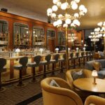 The view of a bar with gray bar stools, wood paneled walls with shelves of alcohol, large chandeliers and small tables with arm chairs.