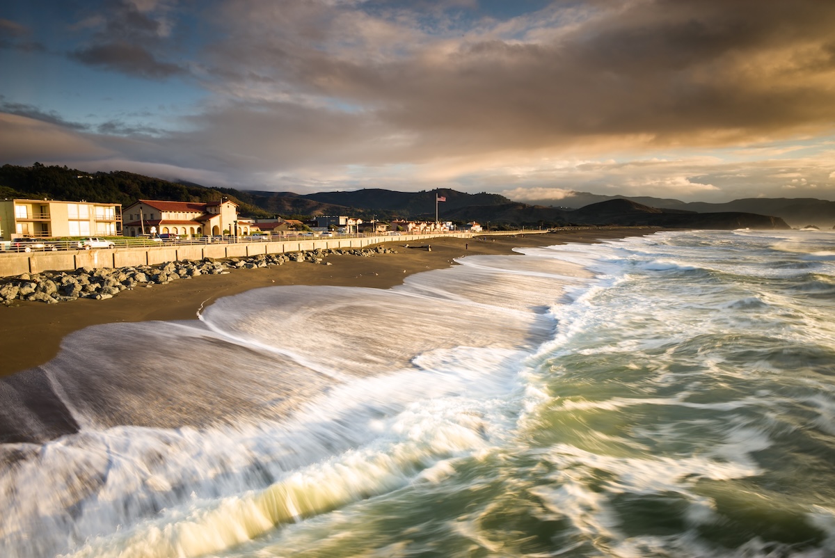 A beach at sunset, with the waves crashing on shore and the road stretching along the edge.