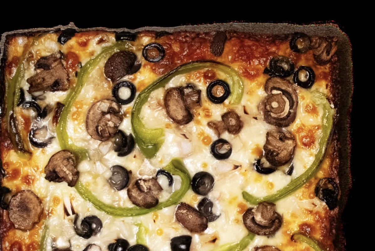 An above shot of a square pizza topped with mushrooms, olives and green bell pepper.