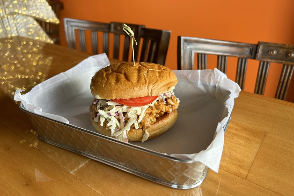 A fried chicken sandwich with a tomato and slaw sits in a tray with a toothpick through the top.