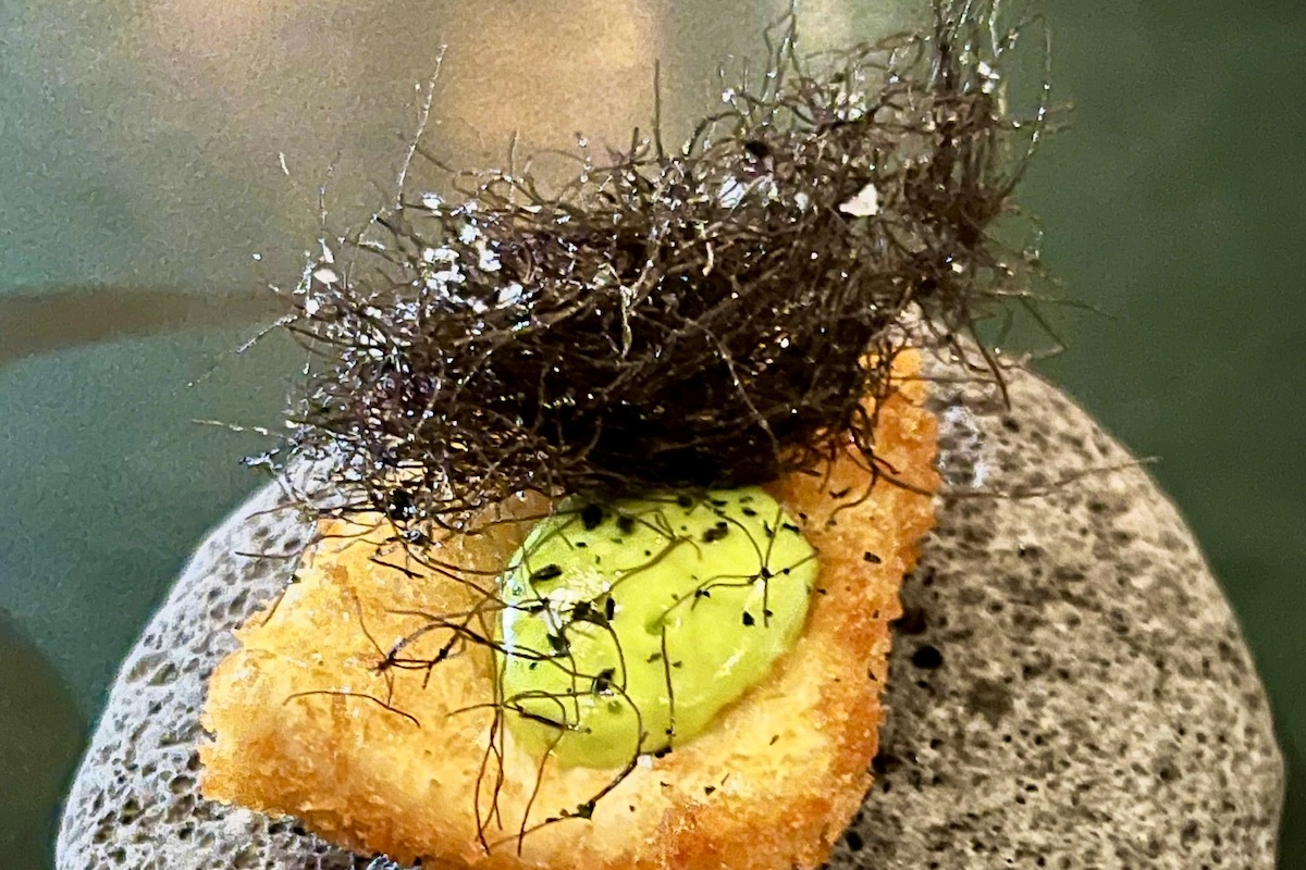 A piece of breaded fish topped with a stringy black topping and dollop of green sauce.