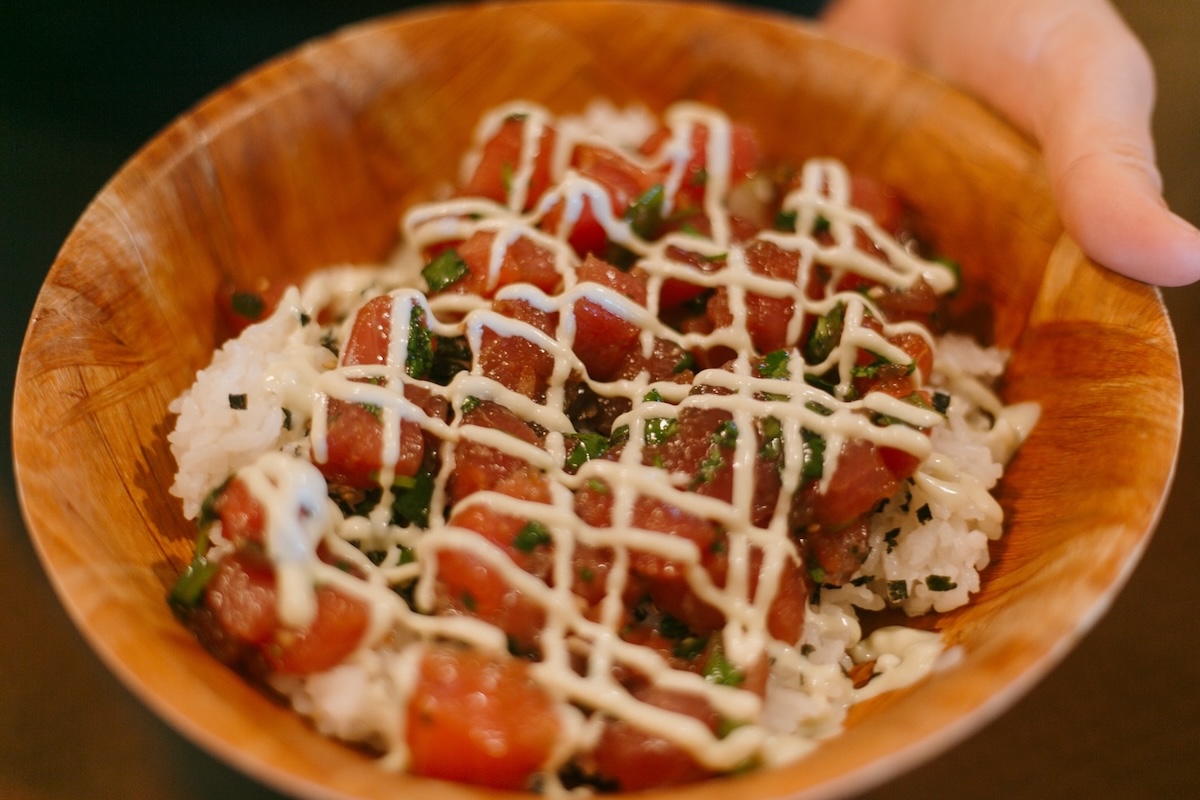 Someone holds a wooden bowl with rice, poke, furikake seasoning and mayo drizzle.