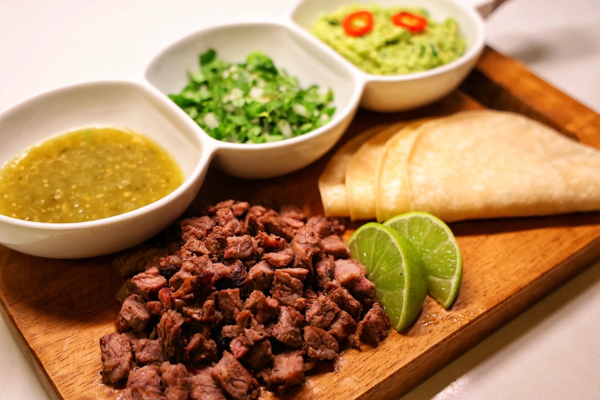 A wooden cutting board with mini corn tortillas and bowls of taco toppings.