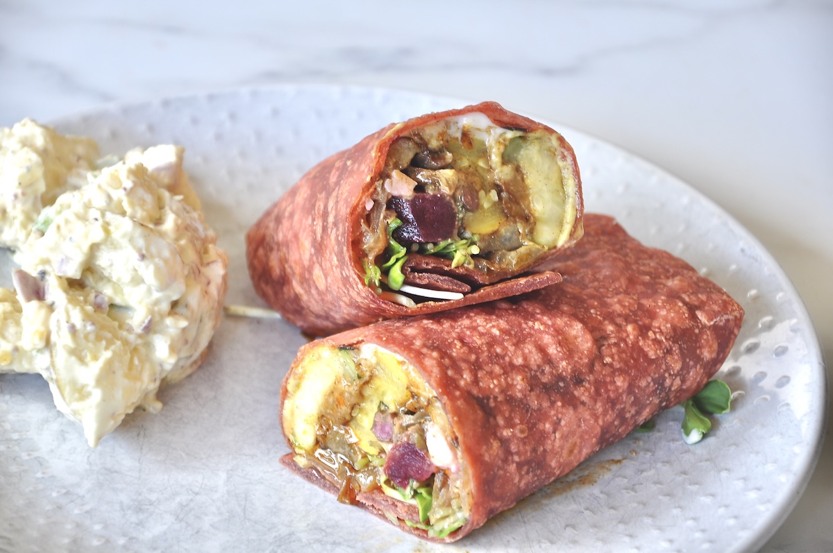 Two halves of a wrap with a beet tortilla and lots of meat and veggies for filling and a scoop if potato salad.
