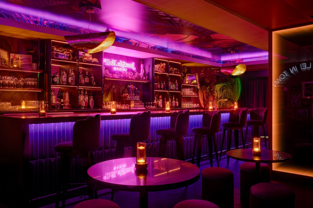 A bar with dimmed lights and bright pink neon light, purple bar stools and stocked alcohol shelves.