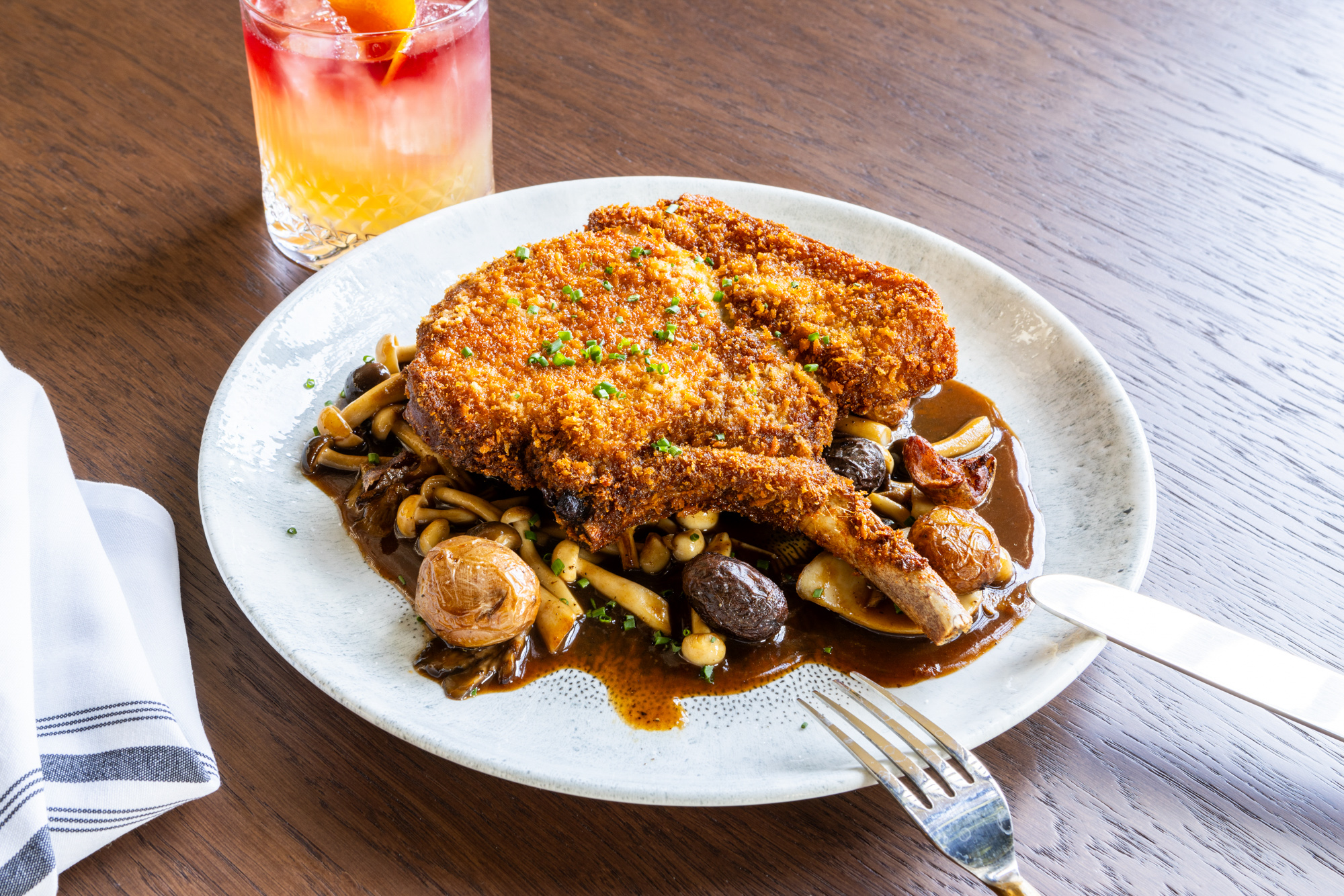 On a plate is breaded meat still on the bone and on a bed of sauce with mini roasted potatoes and mushrooms.