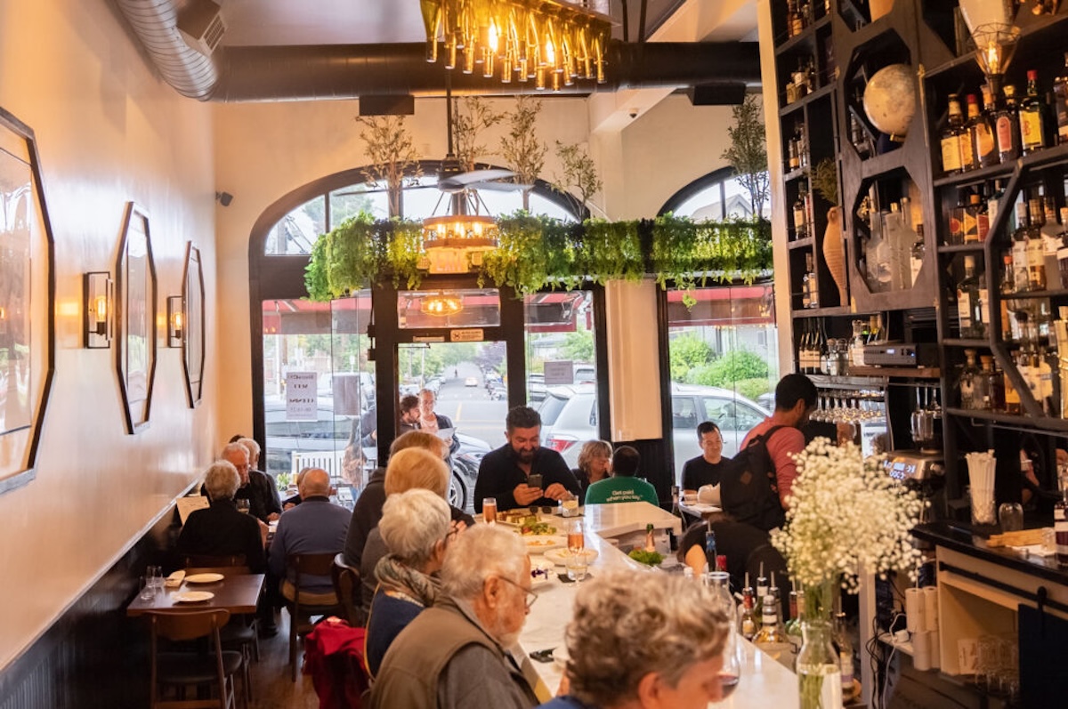 A restaurant with lots of people seated, a chandelier and a bar.