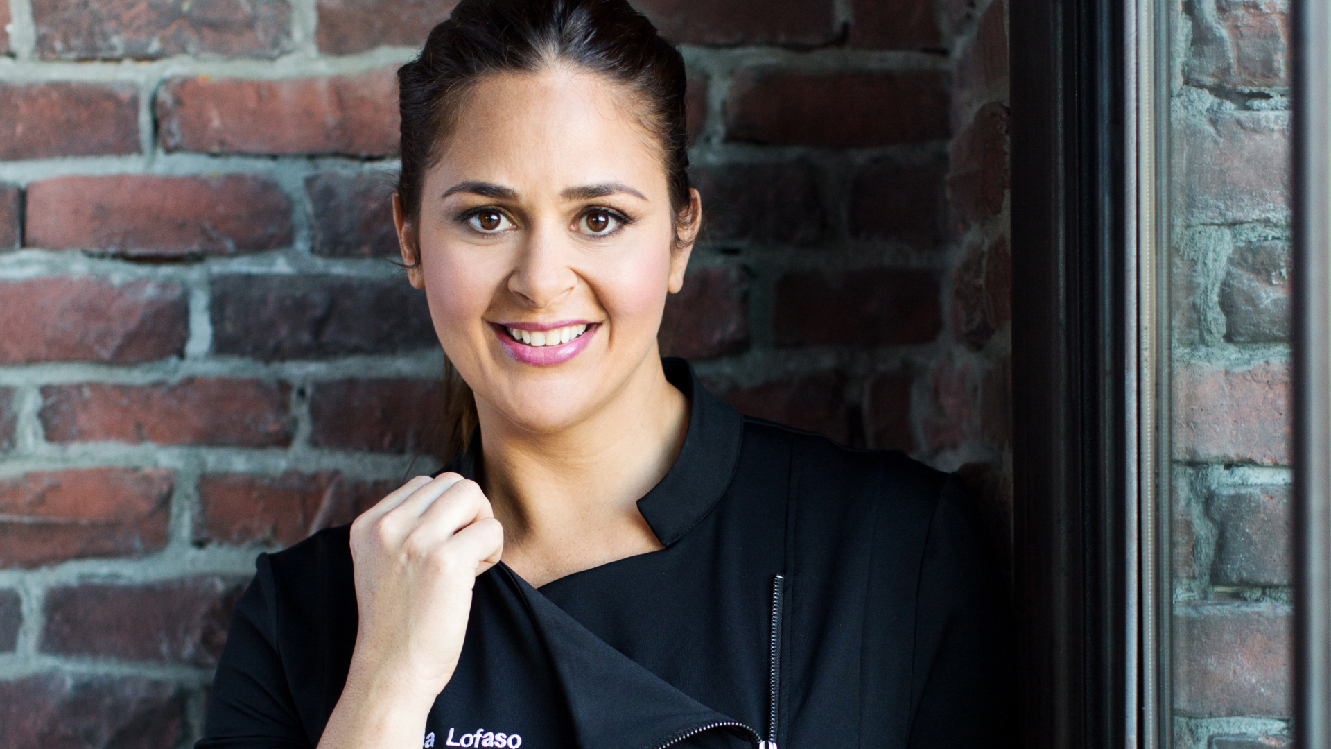Smiling woman in a black chef's coat standing against a brick wall, gently holding the collar with one hand. She has dark hair tied back and is looking directly at the camera.