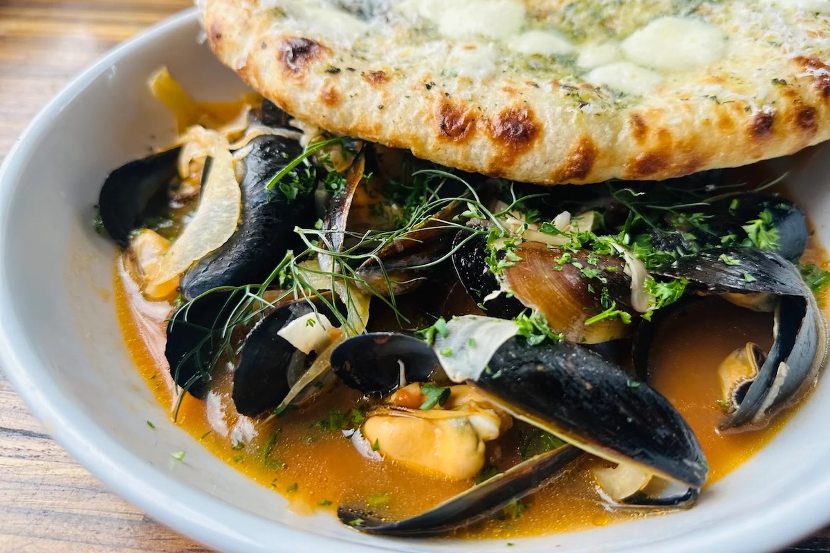 A plate with mussels and shellfish in a sauce and topped with herbs and with a side of bread.