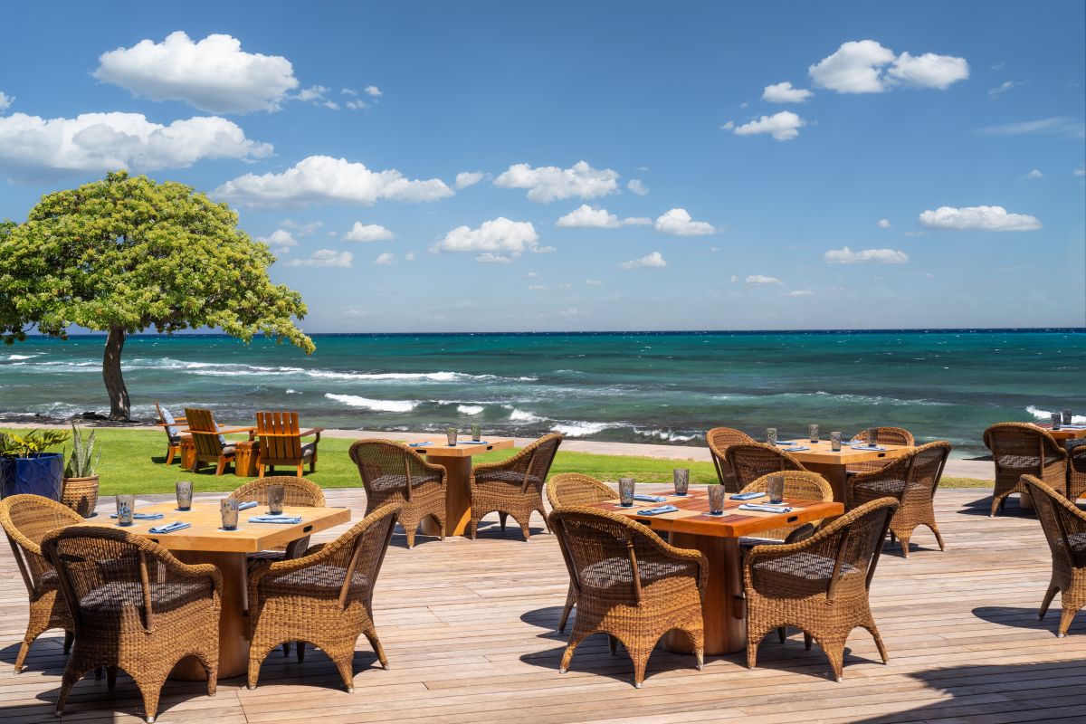 Outdoor dining area with wooden tables and wicker chairs overlooking a scenic ocean view.