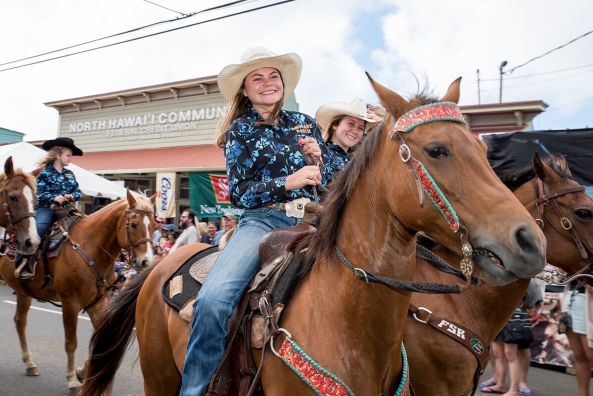 A girl wearing a long sleeve patterned shirt, jeans and a cowboy hat smiles as she rides a brown horse down the street and other ride on horseback around her.