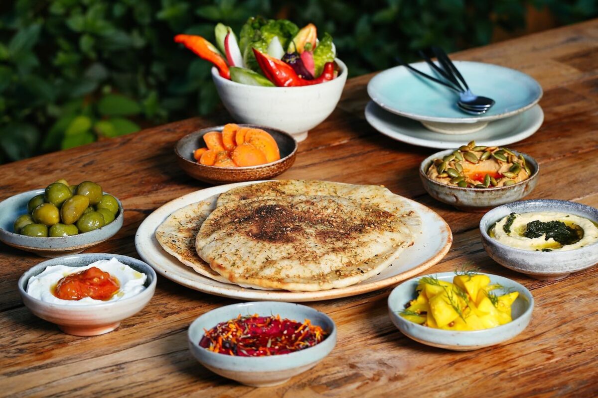 Centered on the table is a plate with pita bread, and there are bowls of beet dip, hummus, fruit, olives, carrots and more around it.