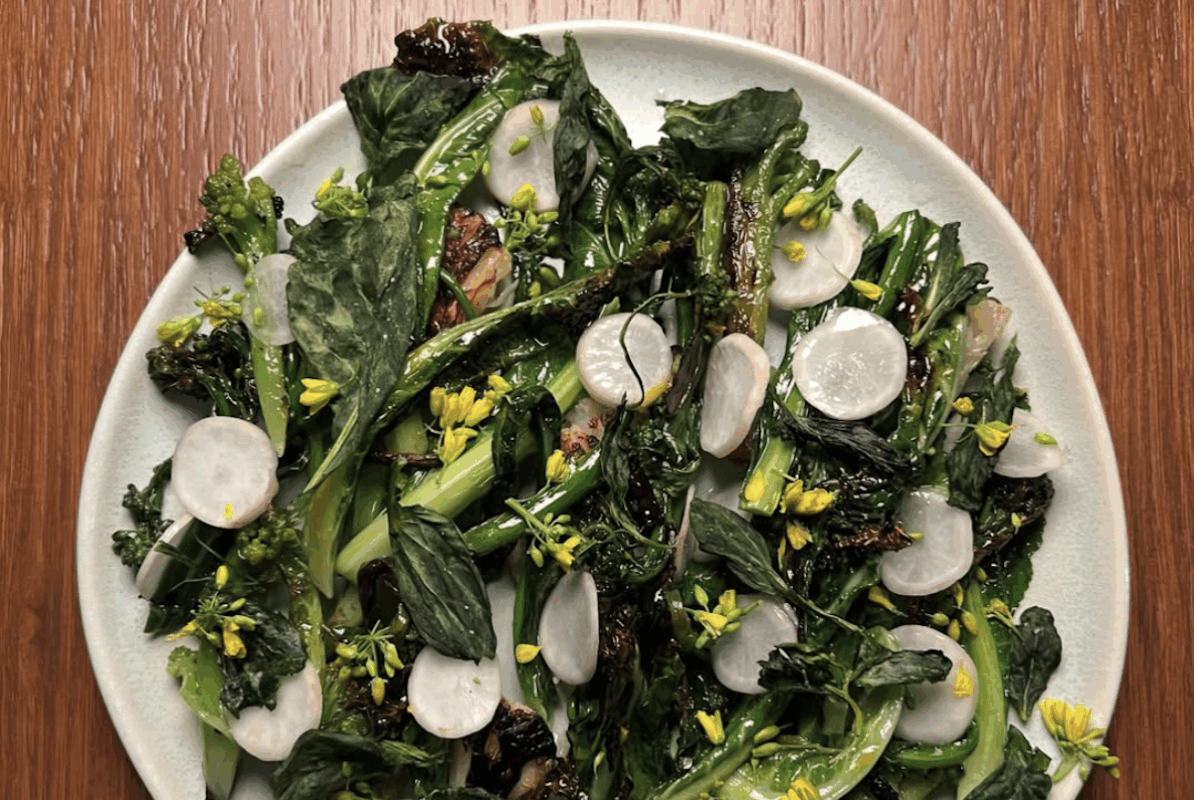 A plate with dark cooked greens and topped with dandelion buds and slices of a round veggie.