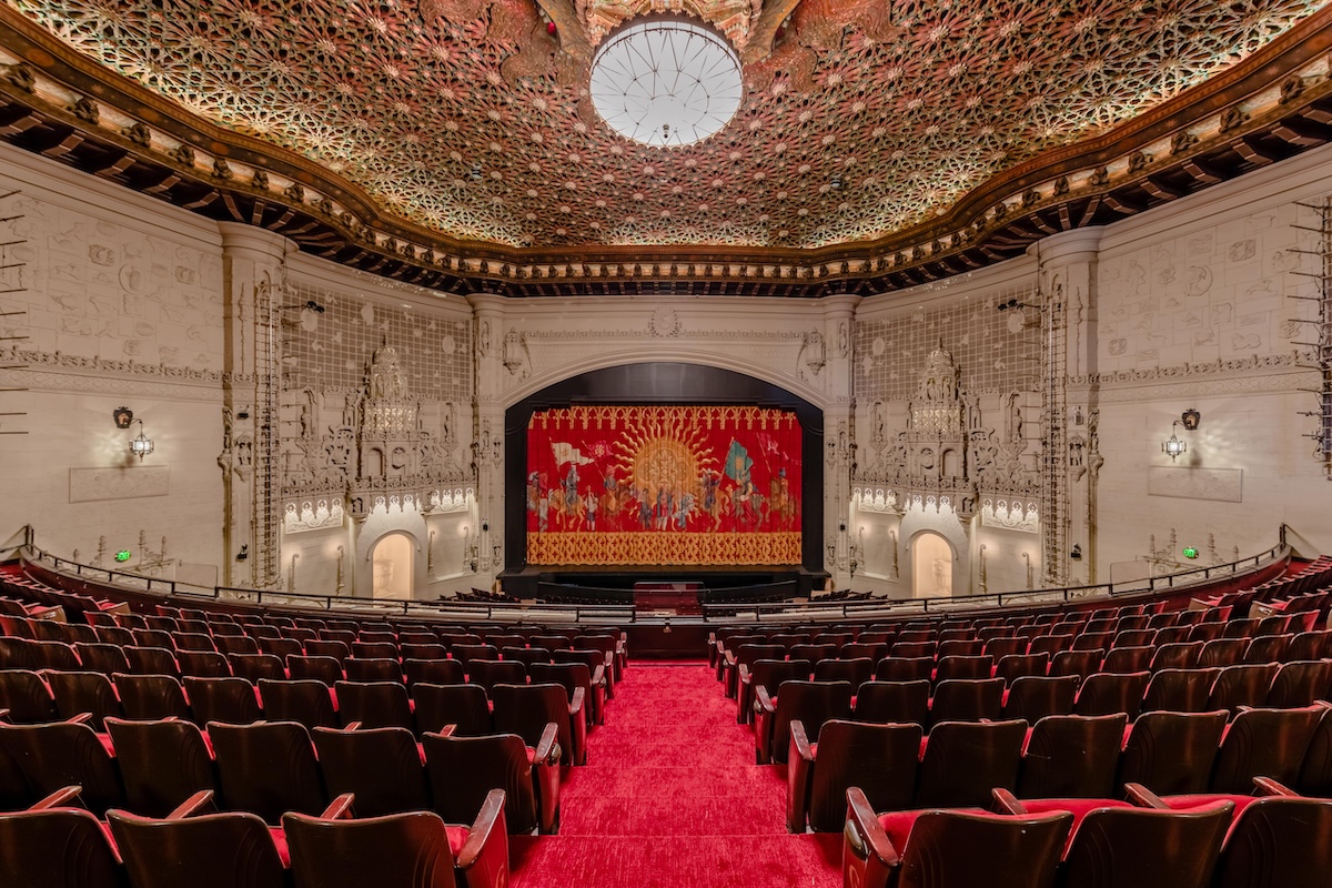 Red theatre seats and carpeting face a stage. The ceiling has an intricate pattern.