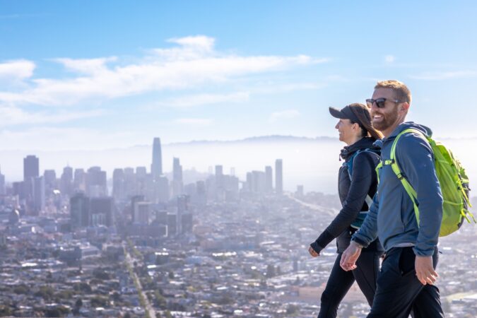 Two hikers in the proper gear and attire walk on a trail with views of San Francisco in the background.