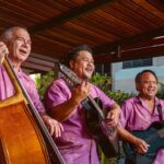 Three men wearing matching fuchsia button down shirts and playing string instruments, including bass and guitar.