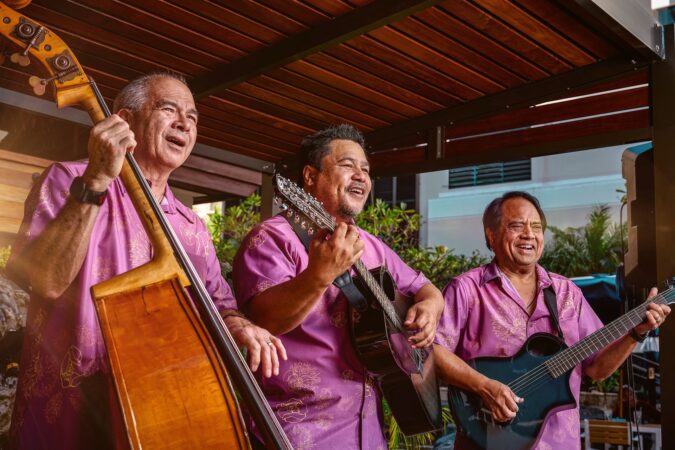 Three men wearing matching fuchsia button down shirts and playing string instruments, including bass and guitar.