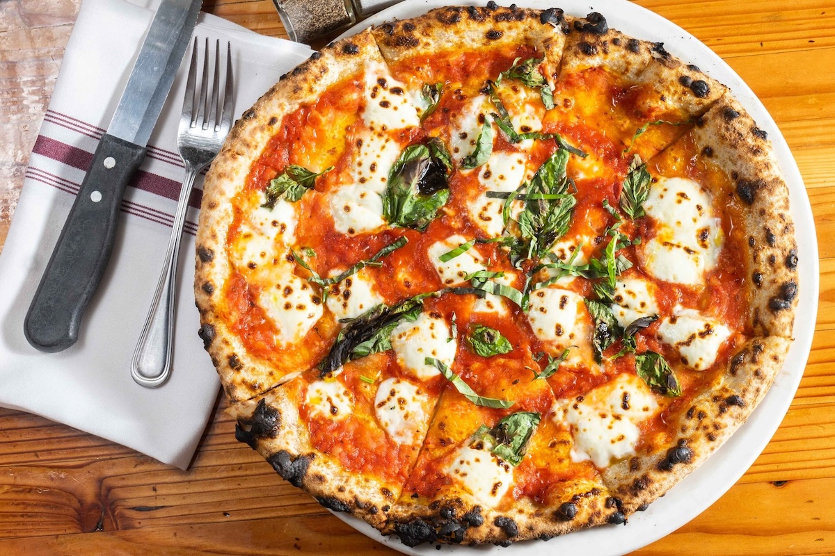 On a wooden table sits a Margherita pizza on a plate with a fork, knife and napkin next to it.
