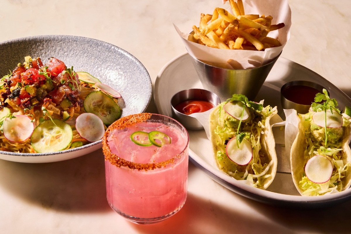 A plate with A noodle salad with veggies and sauce, tacos filled with lettuce and a side of fries and a pink drink.