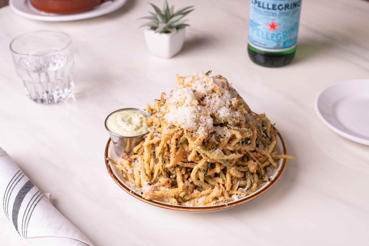 A plate of pasta topped with grated cheese and a side of sauce sits on a table with a glass of water, a bottle on San Pellegrino, a plant and more table settings.