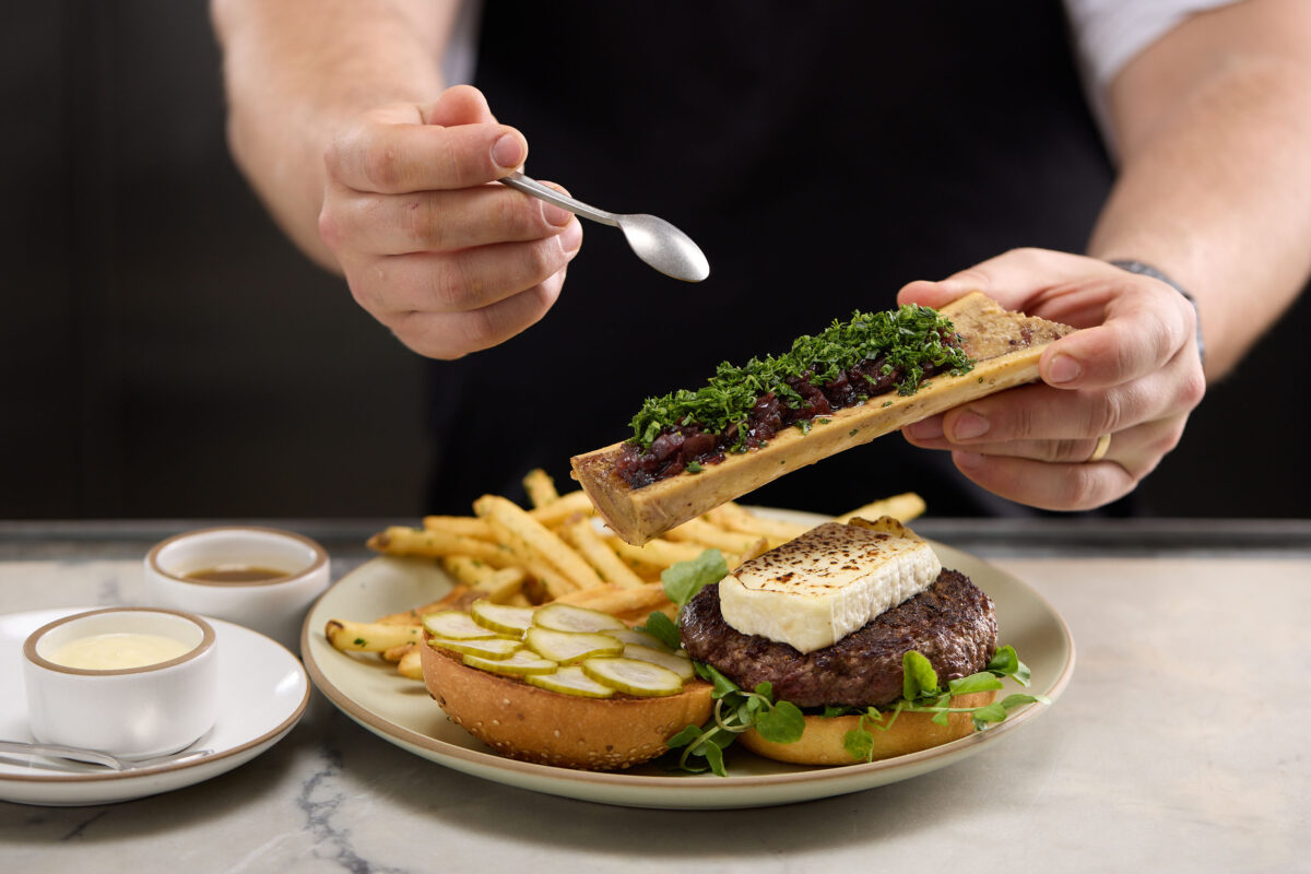 A chef holds a spoon and a bone with bone marrow above a plate with an open faced burger, fries and side sauces.